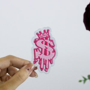 May include: Pink embroidered iron-on patch of a dollar sign with a crown on top. The dollar sign is dripping like melting wax.