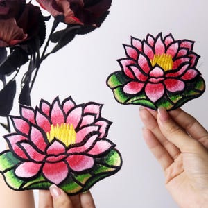 Może przedstawiać: Dwa haftowane łaty z kwiatami lotosu w kolorze różowym i zielonym. Kwiaty mają żółte środki i są otoczone zielonymi liśćmi.