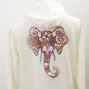 Puede incluir: Sudadera con capucha color crema con un diseño de elefante colorido en la espalda. El elefante está intrincadamente detallado con lentejuelas y bordados en tonos rojos, morados, dorados y marrones. La sudadera tiene capucha y mangas largas.
