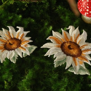 Op de afbeelding: Twee witte geborduurde zonnebloem-patches met bruine centra. De patches bevinden zich op een groen mossig oppervlak.