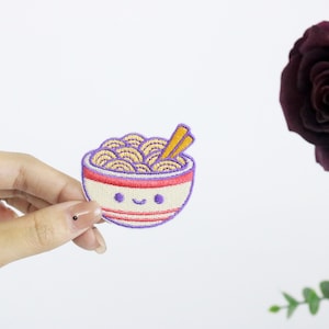 Op de afbeelding: Een witte kom met paarse en rode strepen en een vrolijk gezicht, gevuld met gele noedels en een paar oranje eetstokjes. De kom is geborduurd op een witte stof.