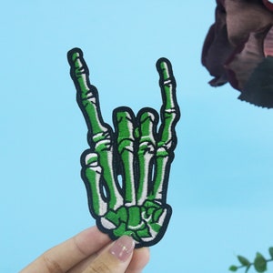 May include: Green skeleton hand making the rock and roll hand gesture. The hand is embroidered on a black fabric patch.