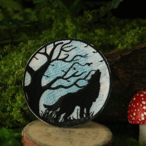 Peut inclure: Un écusson brodé blanc avec une silhouette noire de loup hurlant à la lune. La lune est bleue et le loup se tient devant un arbre aux branches nues.