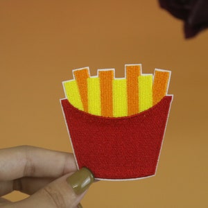 May include: A red and yellow embroidered iron-on patch of a cartoon french fry container with fries.