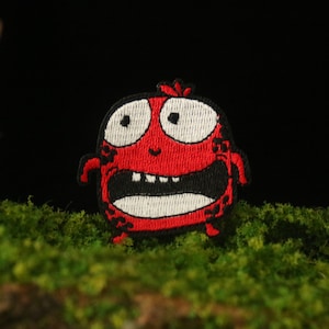 May include: Red embroidered patch of a cartoon monster with white eyes, a wide open mouth with teeth, and black outlines. The monster is on a green mossy surface.