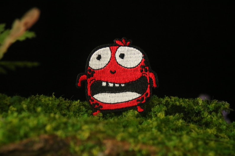 以下が含まれることがあります： 白い目で、歯の生えた大きく開いた口、黒い輪郭を持つ、漫画のモンスターの赤い刺繍パッチ。モンスターは緑の苔むした表面の上にあります。