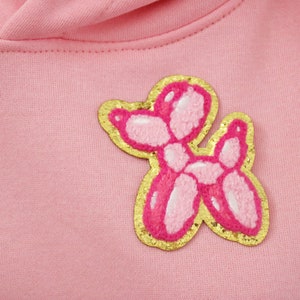 Może przedstawiać: Różowa bluza z kapturem z naszywką w kształcie psa z nadmuchiwanego zwierzęcia w kolorze złotym i różowym.