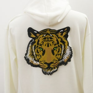 Op de afbeelding: Witte hoodie met een grote geborduurde tijgerkop patch op de rug. De tijger is bruin en zwart met groene ogen.