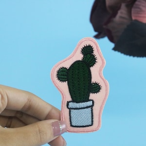 May include: A pink felt cactus patch with green embroidery. The cactus is in a white pot with a grey rim.