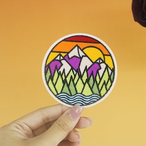 May include: A colorful embroidered patch depicting a mountain range with a sunset behind it. The mountains are purple and white, the sunset is orange and yellow, and the foreground is green trees and a blue river.