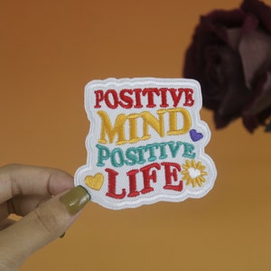 Pode incluir: Etiqueta branca bordada com o texto "POSITIVE MIND POSITIVE LIFE" em letras coloridas. A etiqueta apresenta um sol amarelo com um coração branco no centro e dois corações vermelhos.