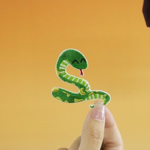 Può includere: Toppa ricamata raffigurante un serpente verde sorridente con la lingua rossa. Il serpente ha dettagli bianchi e gialli, su sfondo arancione. Adatta per abbigliamento o accessori. Misura circa 6 cm di lunghezza.