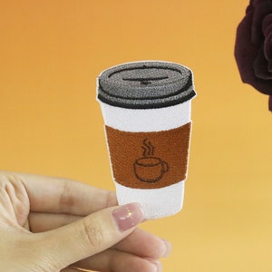 May include: An embroidered patch depicting a white coffee cup with a brown sleeve and a steam design. The cup has a black lid and a small coffee cup icon on the sleeve.