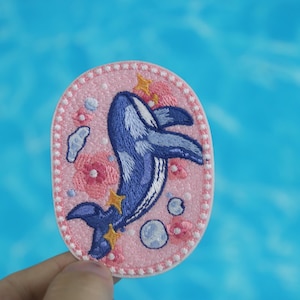 May include: A pink and blue embroidered patch featuring a whale jumping out of the water with stars and bubbles. The patch is oval shaped and has a white border.