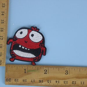 以下が含まれることがあります： 大きく開いた口と大きな目をした漫画のキャラクターの赤い刺繍アイロン接着パッチ。キャラクターの高さは約5cmです。