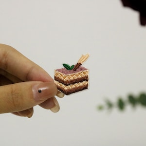 May include: An embroidered patch of a layered cake, featuring brown and beige colours, with a green leaf and a brown stick on top.