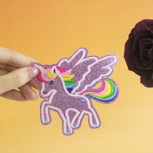 Puede incluir: Un unicornio rosa con purpurina con alas y cola arcoíris. El unicornio está bordado y tiene un acabado brillante.