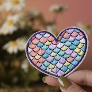 May include: Embroidered heart-shaped patch with a mermaid scale design. The patch features a purple border and a pattern of pastel colors including pink, blue, yellow, and lavender. The patch is likely for clothing or accessories.