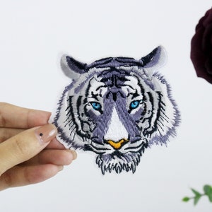 May include: Embroidered iron-on patch of a white tiger's face with blue eyes. The tiger has a gray and white striped pattern.