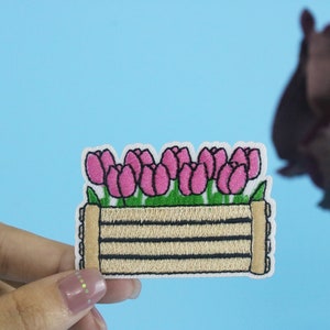 May include: A pink and green embroidered patch of a wooden planter with pink tulips.