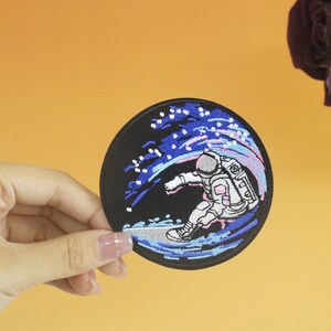 Könnte beinhalten: Ein gestickter Aufnäher mit einem Astronauten, der auf einer Welle im Weltraum reitet. Der Astronaut trägt einen weißen Raumanzug mit einem blauen Helm und hält ein Surfbrett. Die Welle ist blau und rosa mit weißen Sternen.