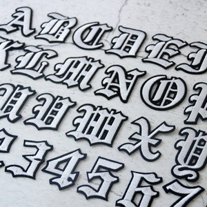 May include: A set of white iron-on letters and numbers in a gothic font style. The letters are outlined in black and have a slightly raised texture.