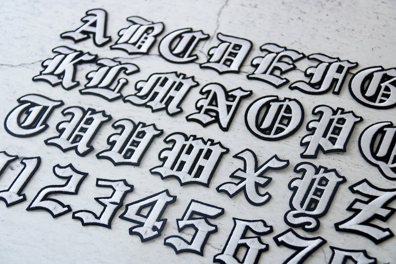 May include: A set of white iron-on letters and numbers in a gothic font style. The letters are outlined in black and have a slightly raised texture.