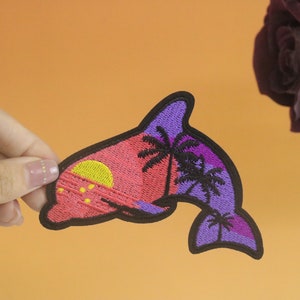 May include: A purple and pink embroidered dolphin patch with a sunset and palm trees. The dolphin is facing right and has a black outline.