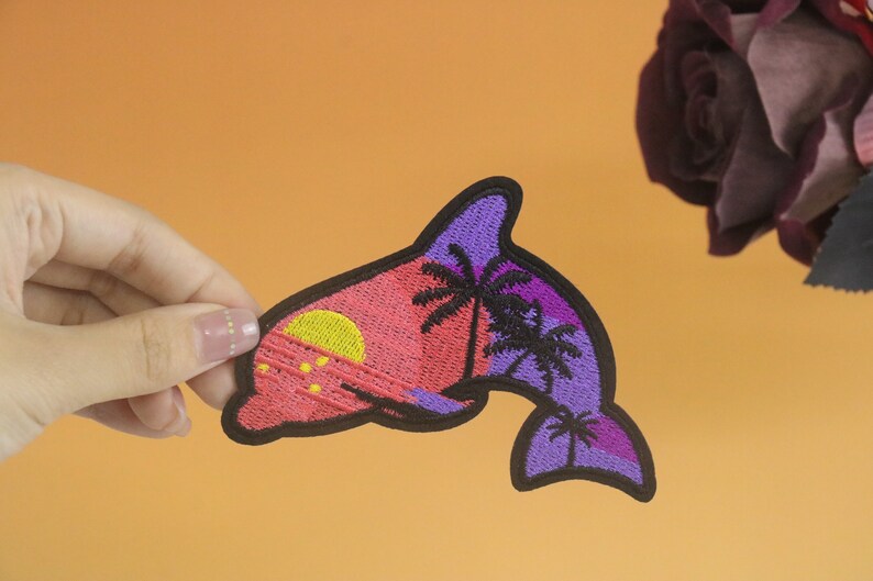 May include: A purple and pink embroidered dolphin patch with a sunset and palm trees. The dolphin is facing right and has a black outline.