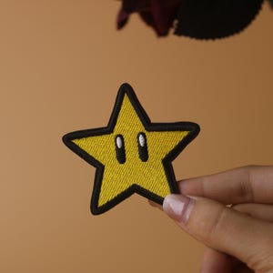 Pode incluir: Um remendo amarelo em forma de estrela com contornos pretos e dois olhos pretos. O remendo é bordado com uma estrela amarela e contornos pretos.