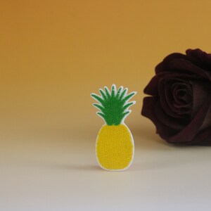May include: A yellow and green pineapple shaped patch with a white background. The pineapple is sitting on a white surface in front of a dark red rose.