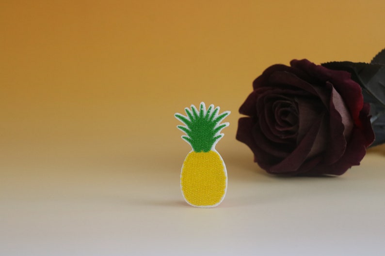 May include: A yellow and green pineapple shaped patch with a white background. The pineapple is sitting on a white surface in front of a dark red rose.