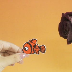 May include: A hand holds a small orange and black embroidered patch of a cartoon clownfish. The fish has a large eye and a small mouth. The patch is on a light orange background.