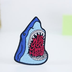以下が含まれることがあります： 青い刺繍の入ったサメのパッチ。口が開いていて、赤い文字で「Wish you were here」と書かれています。