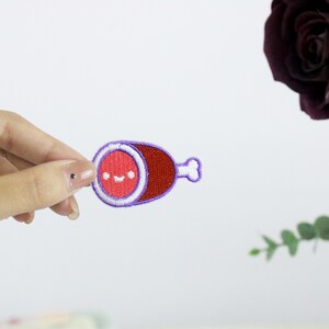 May include: A small, embroidered patch shaped like a ham with a smiling face. The ham is red and the outline is purple.