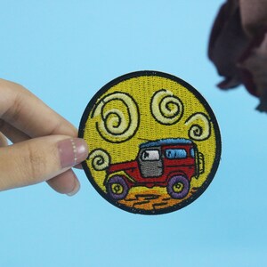 May include: A yellow embroidered patch with a red jeep driving through a yellow sun with white swirls. The jeep has a blue roof and black wheels.