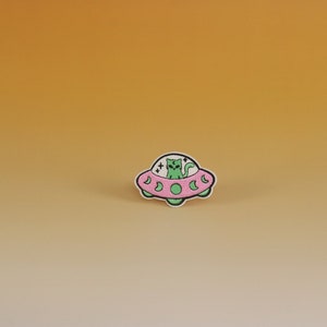May include: An embroidered patch featuring a green cat riding in a UFO. The UFO is pink with white accents. The cat has a white face and black eyes.