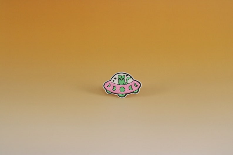 May include: An embroidered patch featuring a green cat riding in a UFO. The UFO is pink with white accents. The cat has a white face and black eyes.