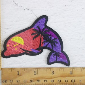 May include: Embroidered patch of a purple dolphin with a sunset and palm trees in the background. The dolphin is outlined in black and the sunset is a gradient of pink, orange, and yellow.