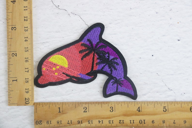 May include: Embroidered patch of a purple dolphin with a sunset and palm trees in the background. The dolphin is outlined in black and the sunset is a gradient of pink, orange, and yellow.