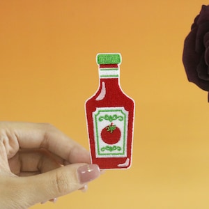 May include: A red and white embroidered ketchup bottle patch with a green lid and a green tomato on the label.