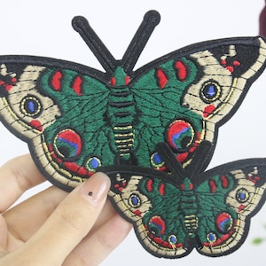 Op de afbeelding: Twee geborduurde strijkpatches met groene, rode en gouden vlinders met zwarte contouren.