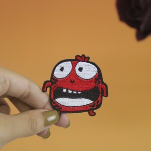以下が含まれることがあります： 白い目と歯を持つ漫画のモンスターの赤い刺繍アイロン接着パッチ。モンスターは驚いた表情をしています。