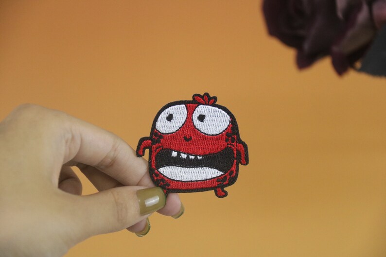 以下が含まれることがあります： 白い目と歯を持つ漫画のモンスターの赤い刺繍アイロン接着パッチ。モンスターは驚いた表情をしています。