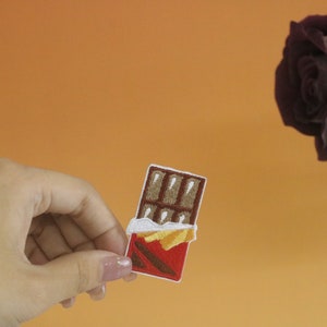 May include: A brown chocolate bar with a red wrapper and yellow foil. The chocolate bar is embroidered on a white fabric patch.