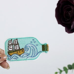 May include: An embroidered patch depicting a ship in a bottle, with waves and bubbles. The patch is blue and brown, and is on a white background.