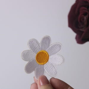 Puede incluir: Un parche de margarita blanca bordada con un centro amarillo. El parche se sostiene en una mano sobre un fondo blanco.