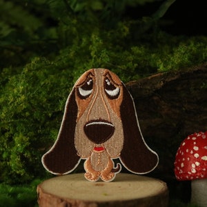 Könnte beinhalten: Gestickter Aufnäher eines braunen und weißen Basset Hounds mit traurigem Gesichtsausdruck. Der Aufnäher befindet sich auf einer Holzoberfläche mit einem Hintergrund aus grünem Moos.