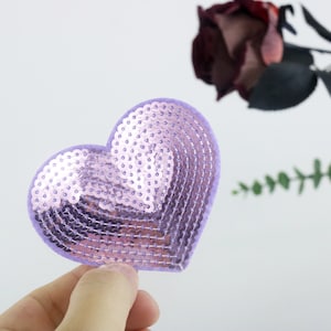 Può includere: Un cerotto a forma di cuore con paillettes viola. Le paillettes sono disposte in un motivo che crea un effetto tridimensionale.