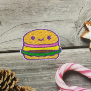 May include: A cute embroidered patch of a smiling hamburger with purple, yellow, pink, green, and brown details.